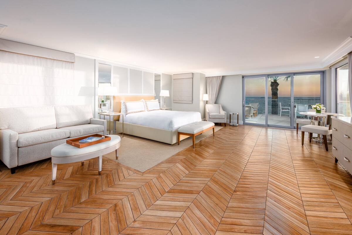 A modern hotel room with a large bed, a sofa, a dining area, and a balcony with a view of the sea.