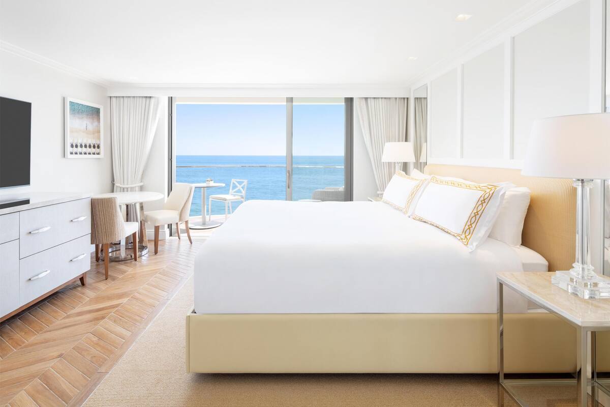 A hotel room with a large bed, a TV, a desk, a chair, and a view of the sea.