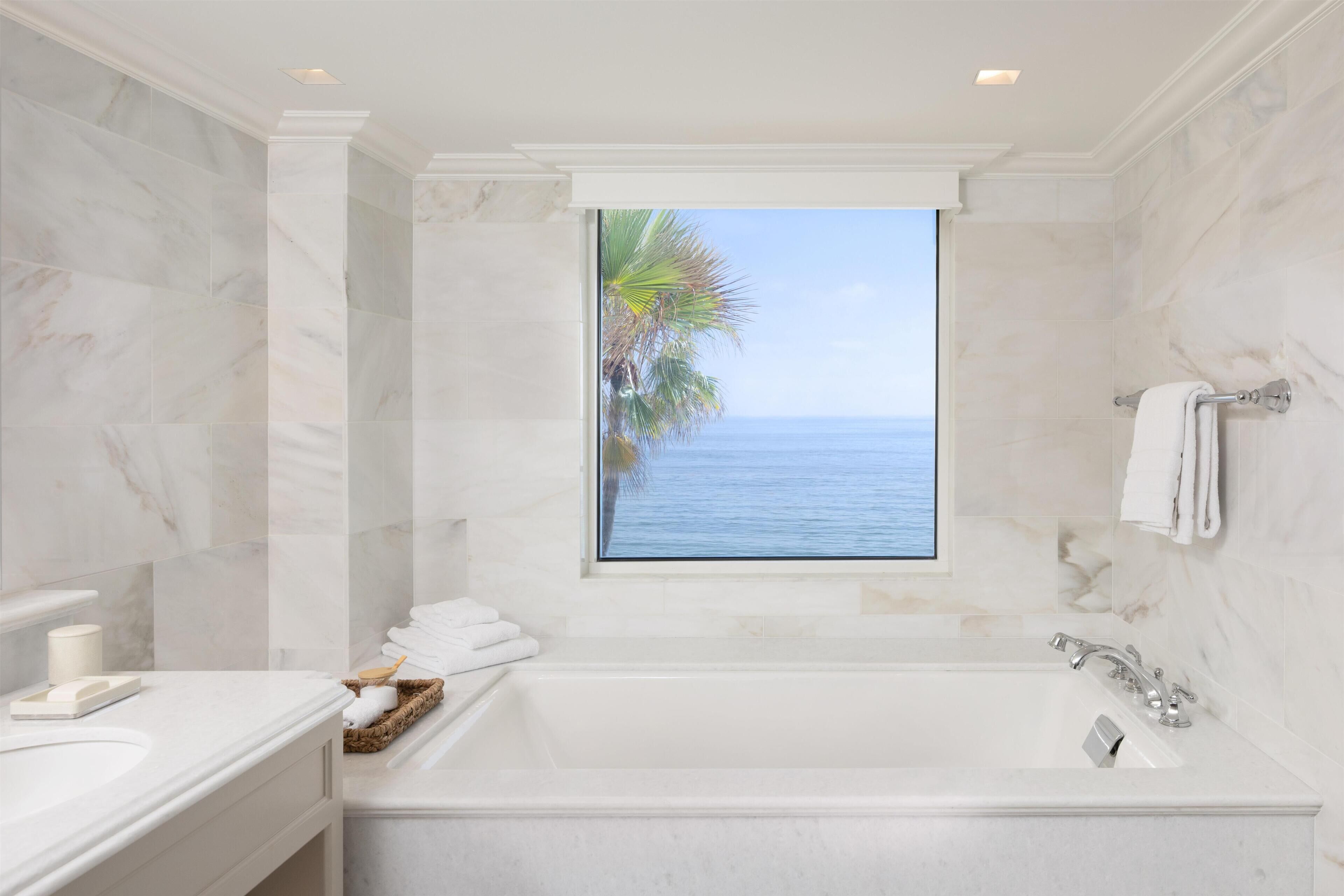 A bathroom with a large bathtub, a window with a view of the sea, and a marble wall.