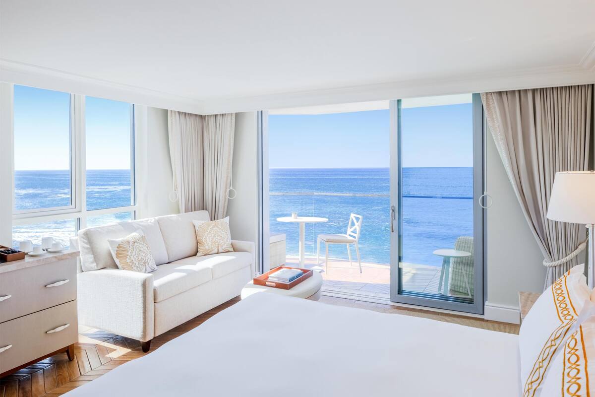 A hotel room with a large bed, a sofa, a small table, and a view of the ocean through large windows.