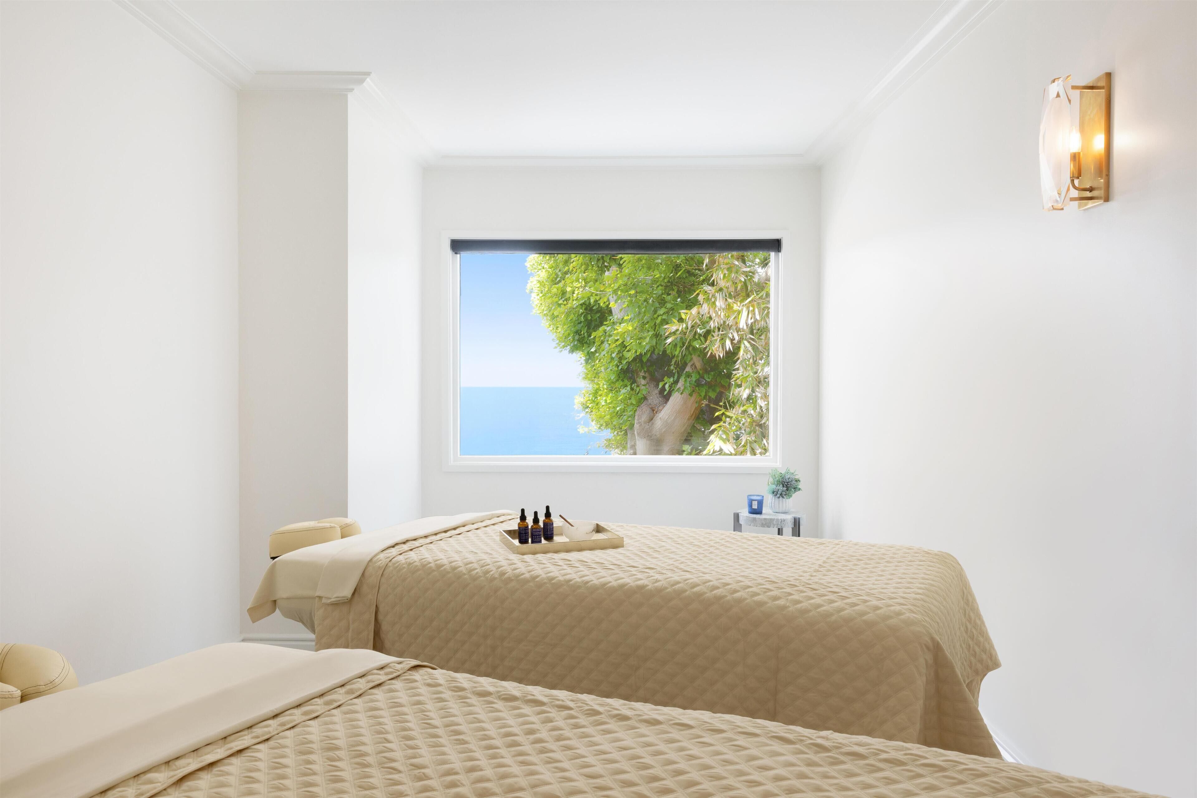 A room with a large window, a bed, a small table with bottles, and a wall-mounted light fixture.