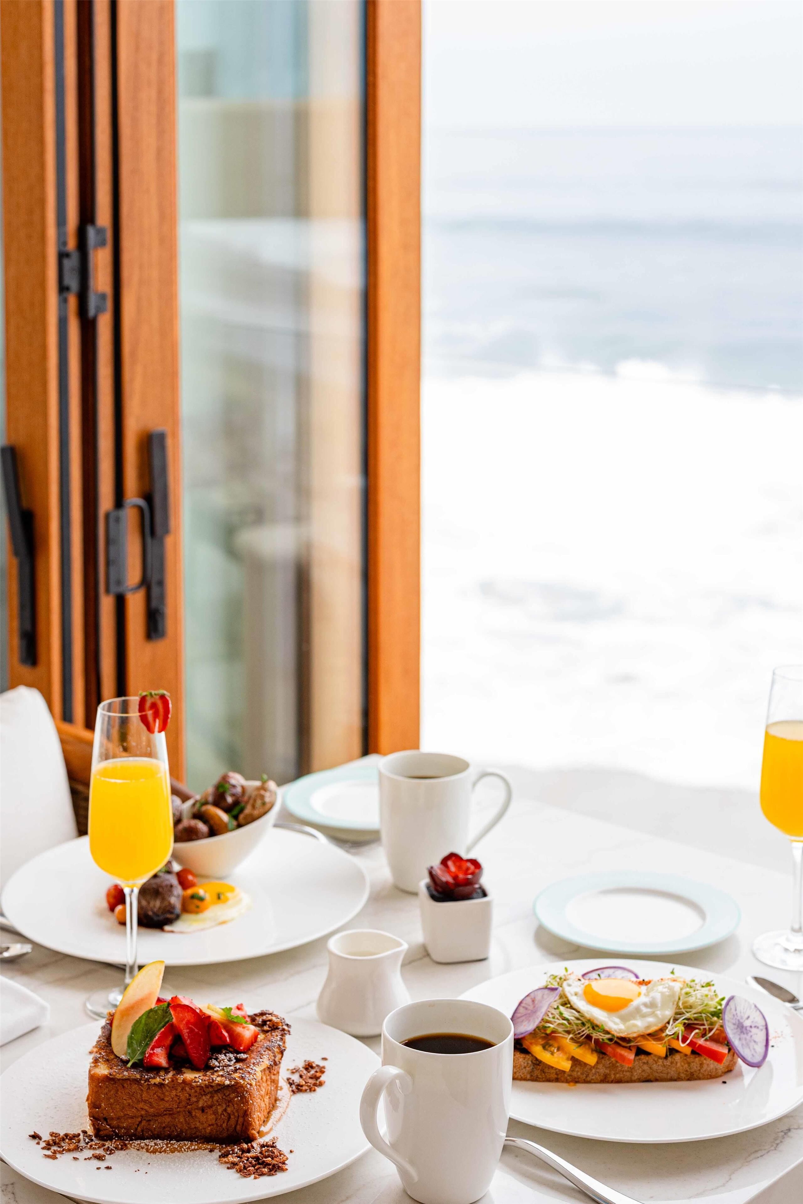 Une table de petit-déjeuner avec vue sur l’océan, comprenant des tartines garnies de fruits, un œuf au plat, un verre de jus d’orange et une tasse de café.