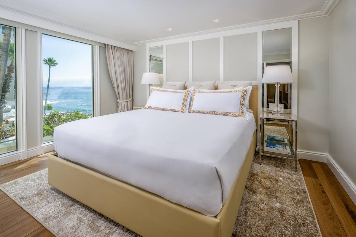 A large bed with a white bedspread and gold trim, a nightstand with a lamp, and a view of the ocean through large windows.