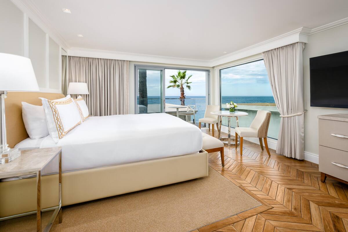 A hotel room with a large bed, a TV, a desk with a lamp, a chair, a small table with a plant, and a view of the ocean.