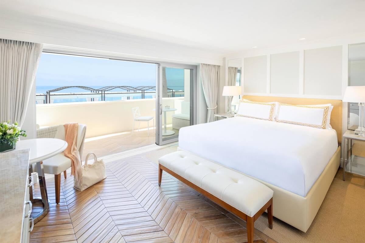 A hotel room with a large bed, a dining table, a bench, and a view of the sea.