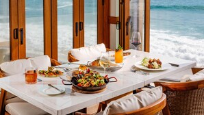 A dining table with seafood dishes, a glass of wine, and a view of the ocean through large wooden doors. - Surf & Sand Laguna Beach (Laguna Beach)