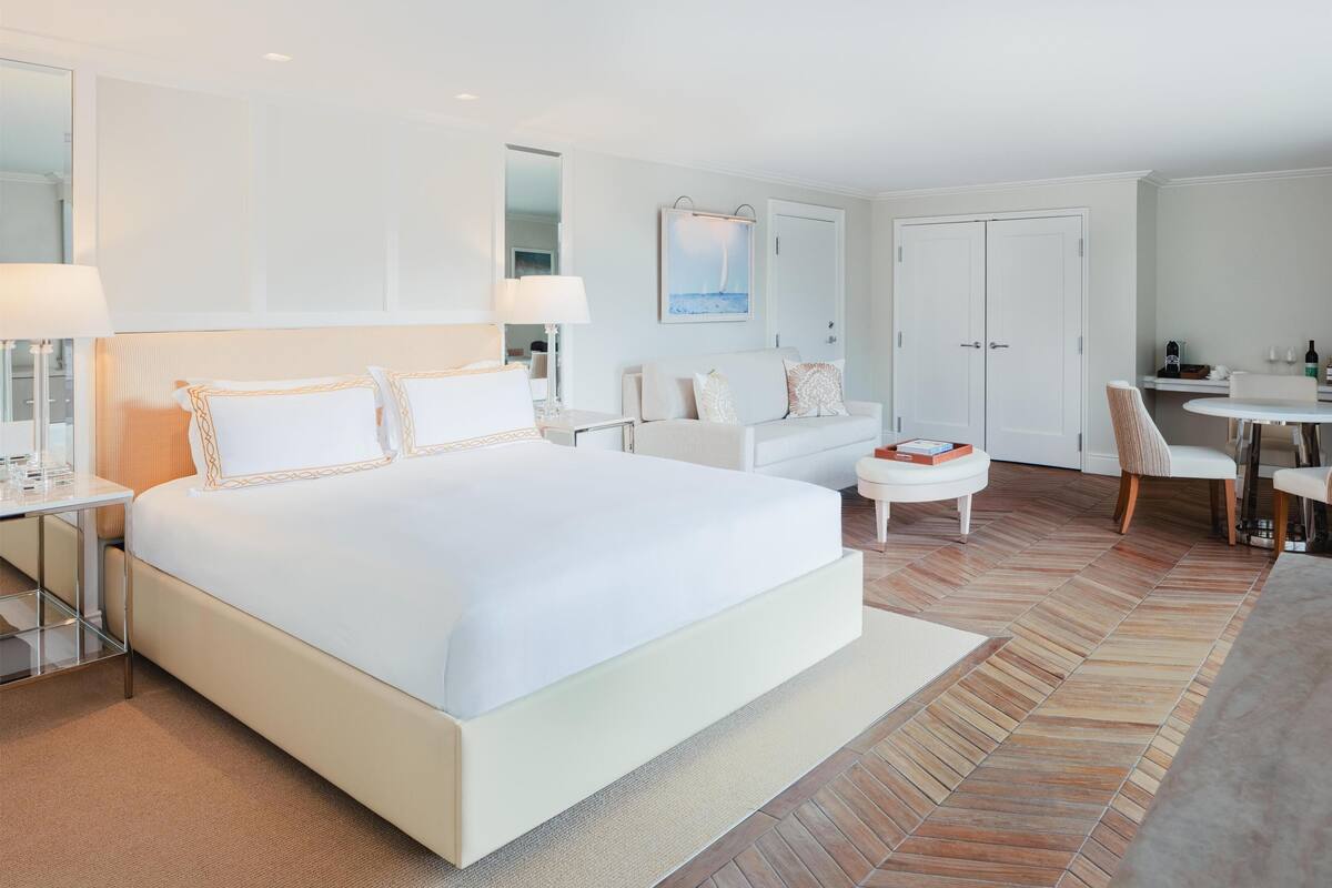 A modern hotel room with a large bed, a sitting area, a dining table, and a separate workspace.