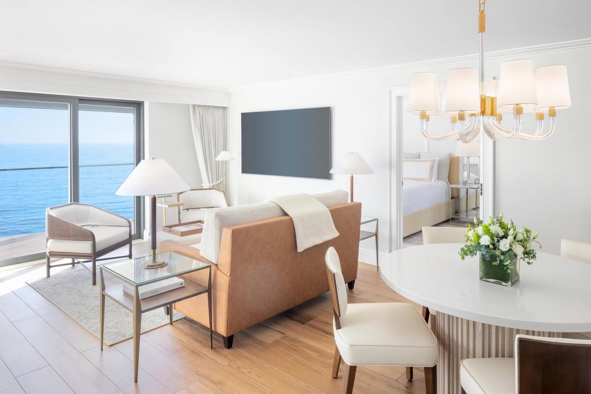 A modern hotel room with a sofa, a dining table, a TV, and a view of the sea.
