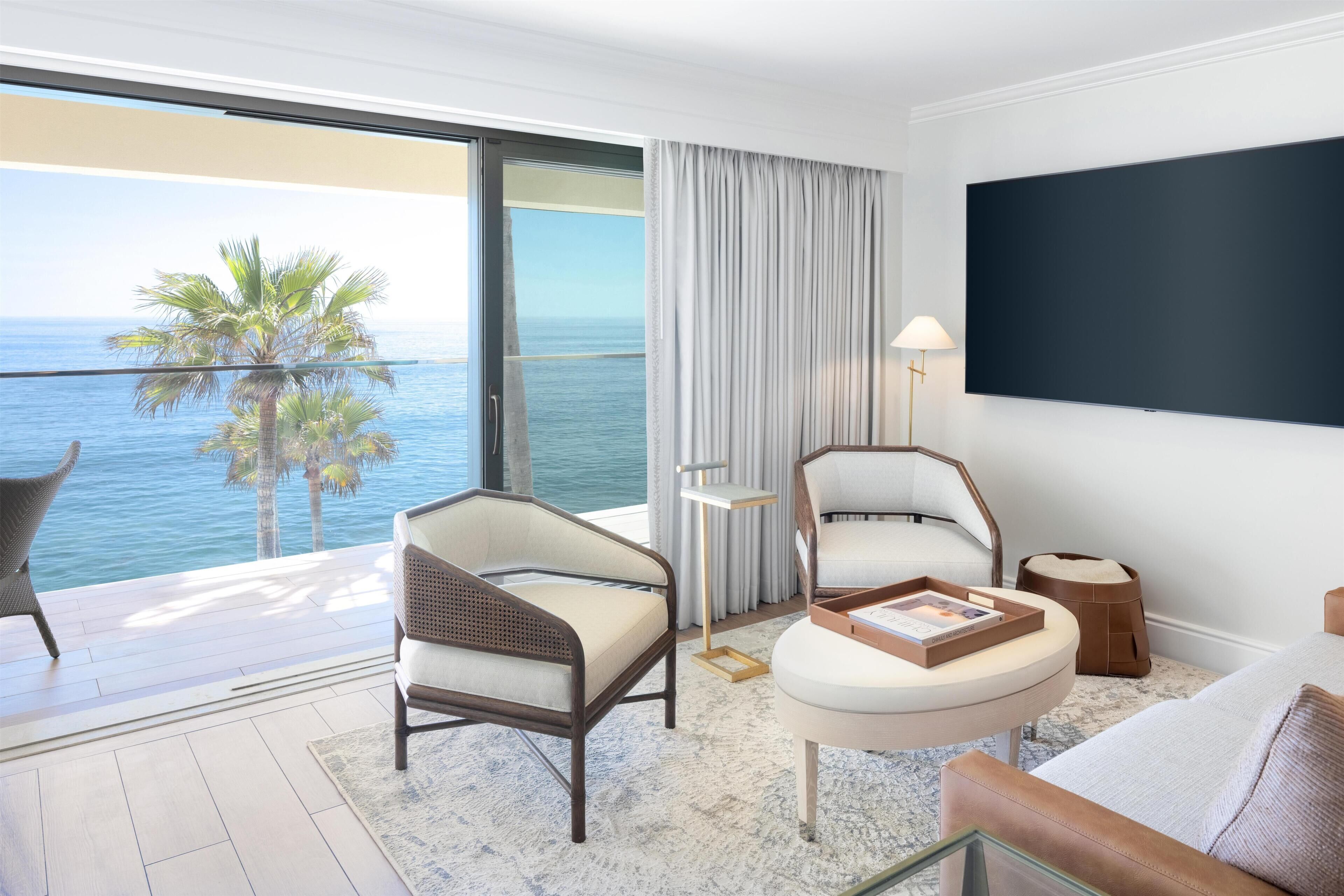 A hotel room with a balcony, a flat-screen TV, two chairs, a small table, and a view of the ocean.