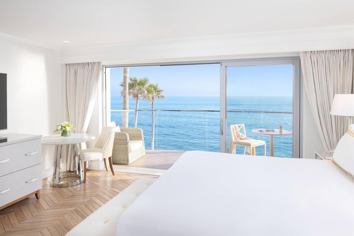 A hotel room with a large bed, a TV, a desk with a chair, a small table with a chair, and a balcony with a view of the sea and palm trees.