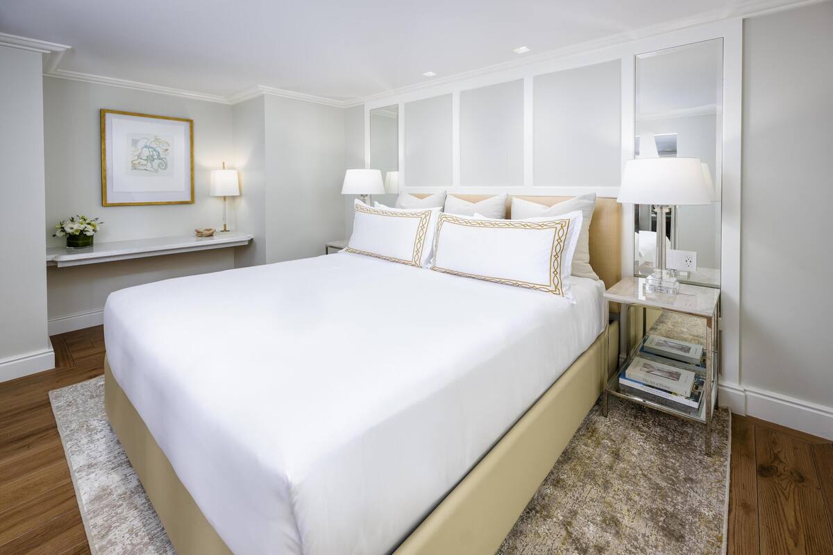 A large bed with white bedding and gold trim, a bedside table with a lamp, a framed picture on the wall, and a small shelf with a vase of flowers.