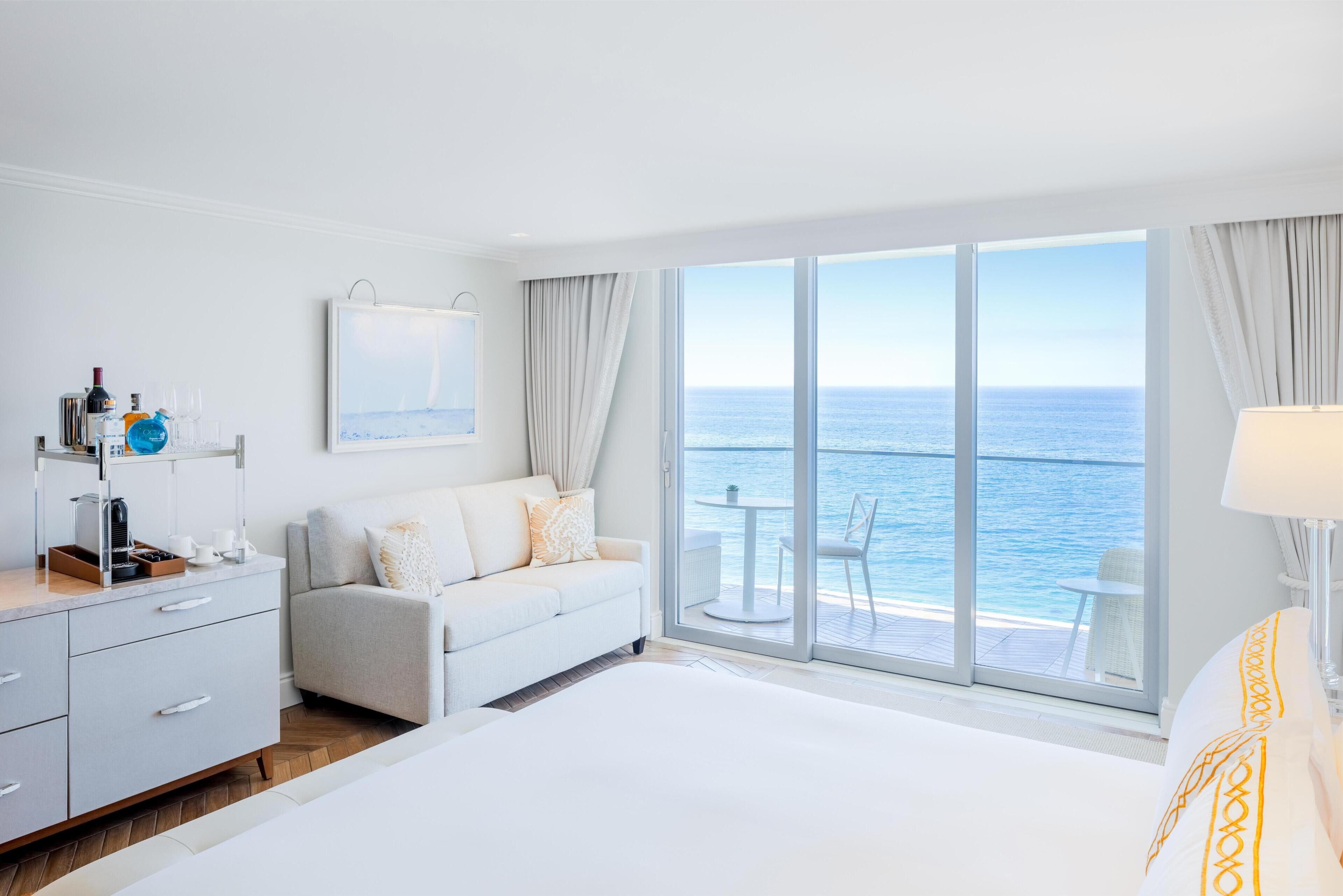 Habitación de hotel con una cama grande, un sofá, una mesa de comedor y vista al océano.