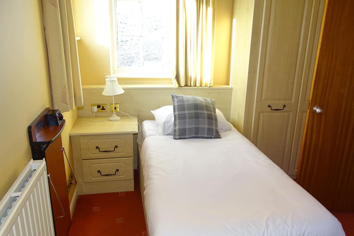 A small, neatly arranged bedroom with a single bed, a bedside table with a lamp, a nightstand, and a wooden door.