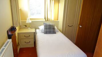 A small, neatly arranged bedroom with a single bed, a bedside table with a lamp, a nightstand, and a wooden door.
