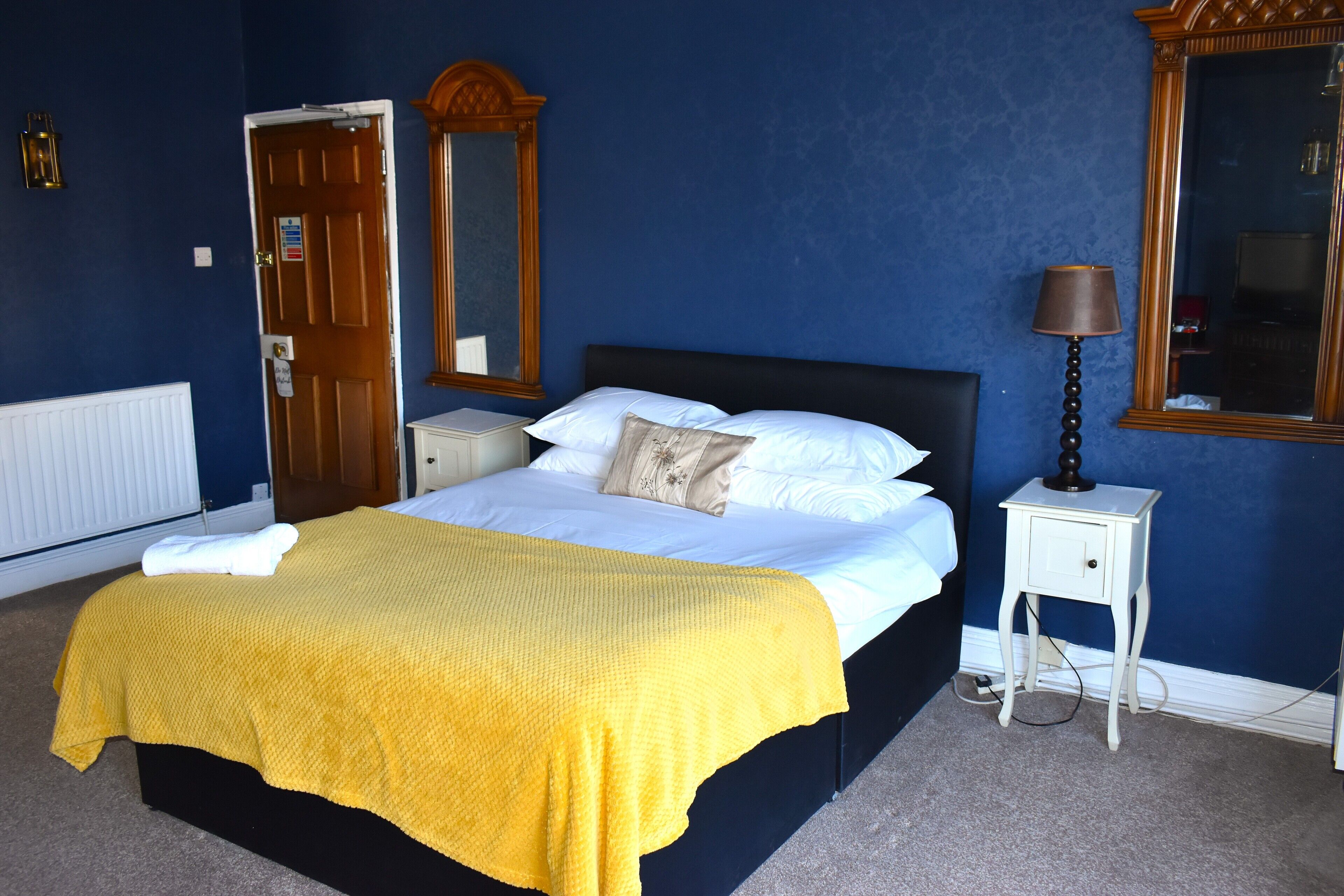 A bedroom with a bed, a bedside table, a lamp, a mirror, and a door.