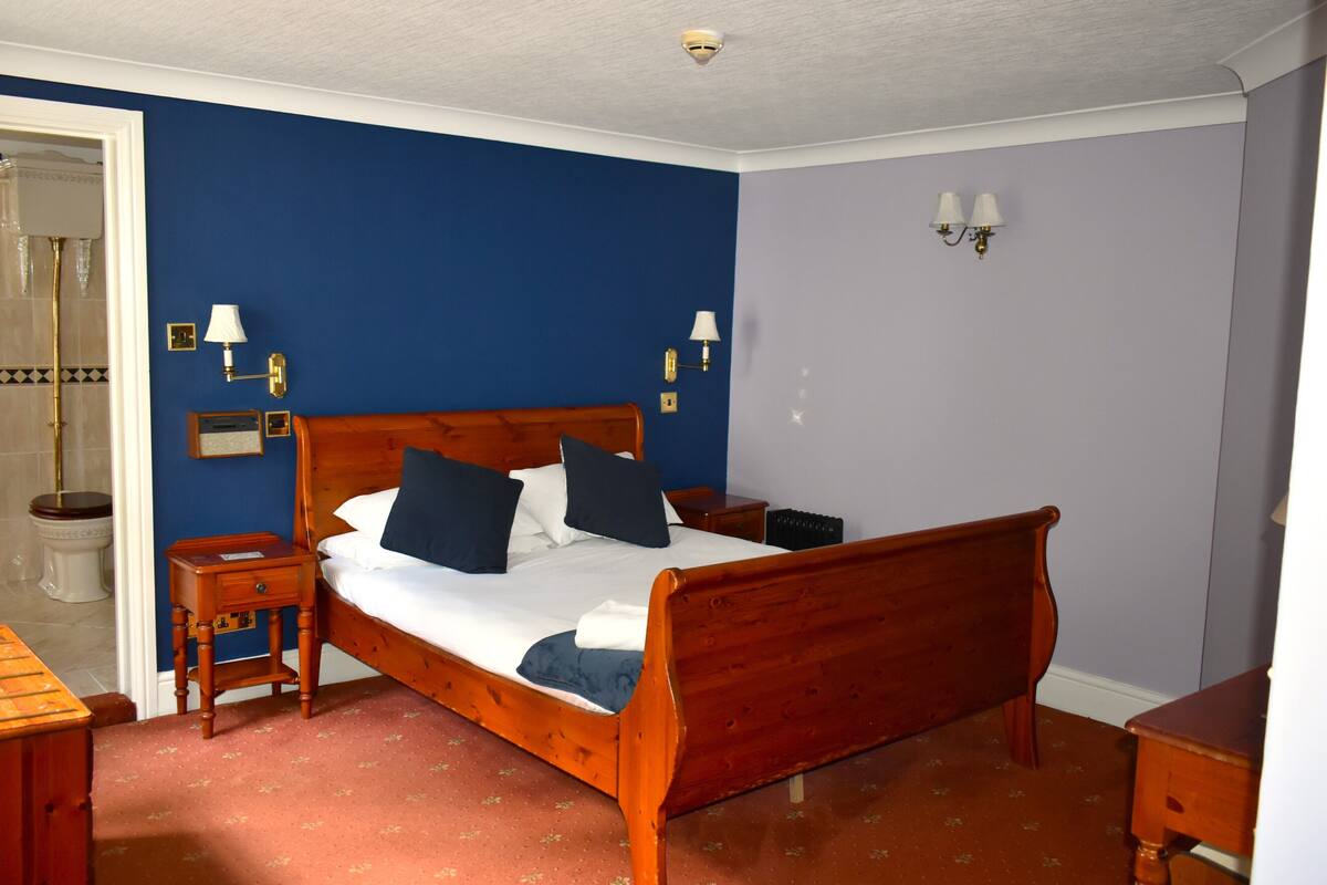 A bedroom with a wooden bed, two bedside tables, a nightstand, a wall-mounted lamp, and a door leading to a bathroom.