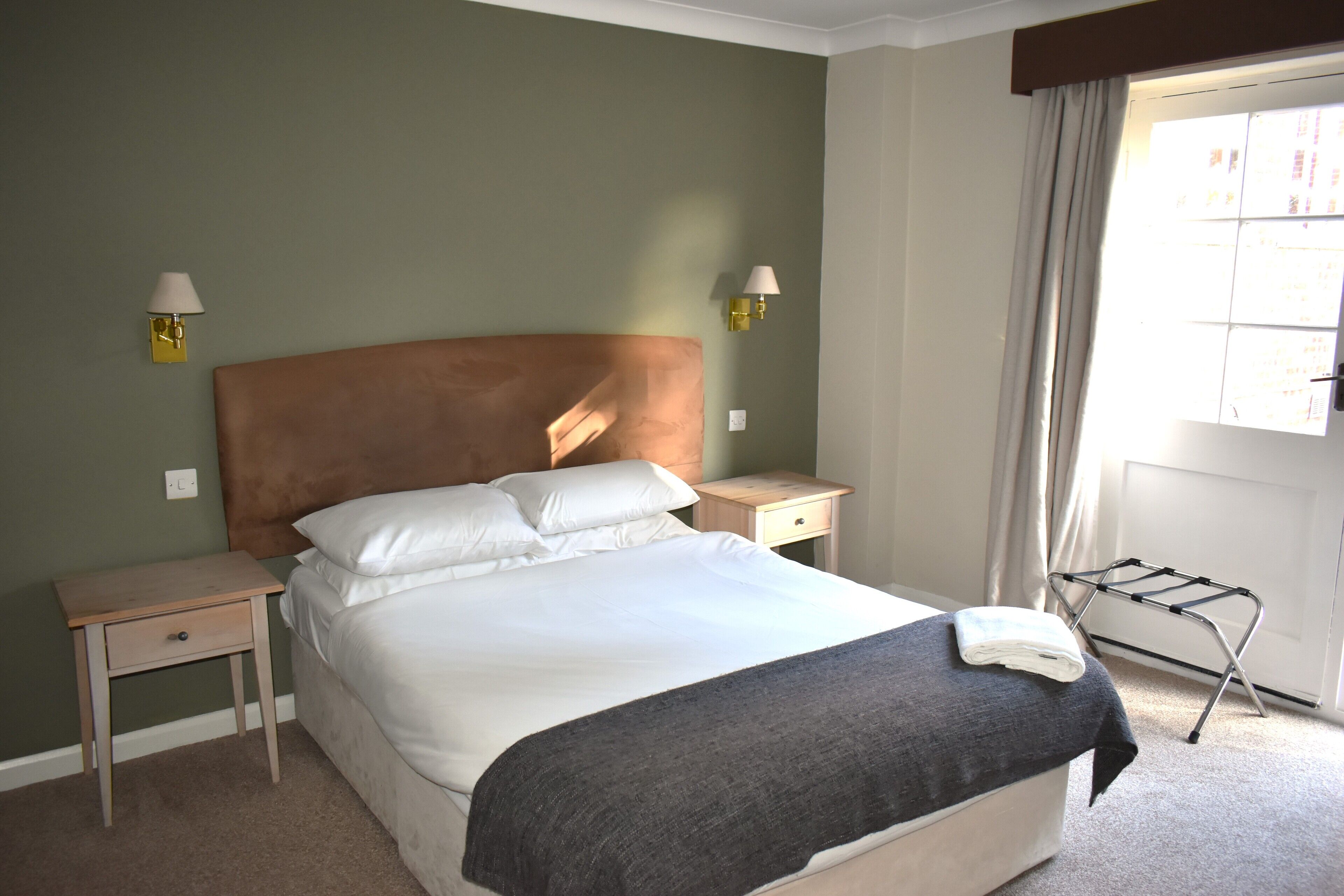A neatly made bed with a headboard, two bedside tables, a lamp, and a window with curtains.