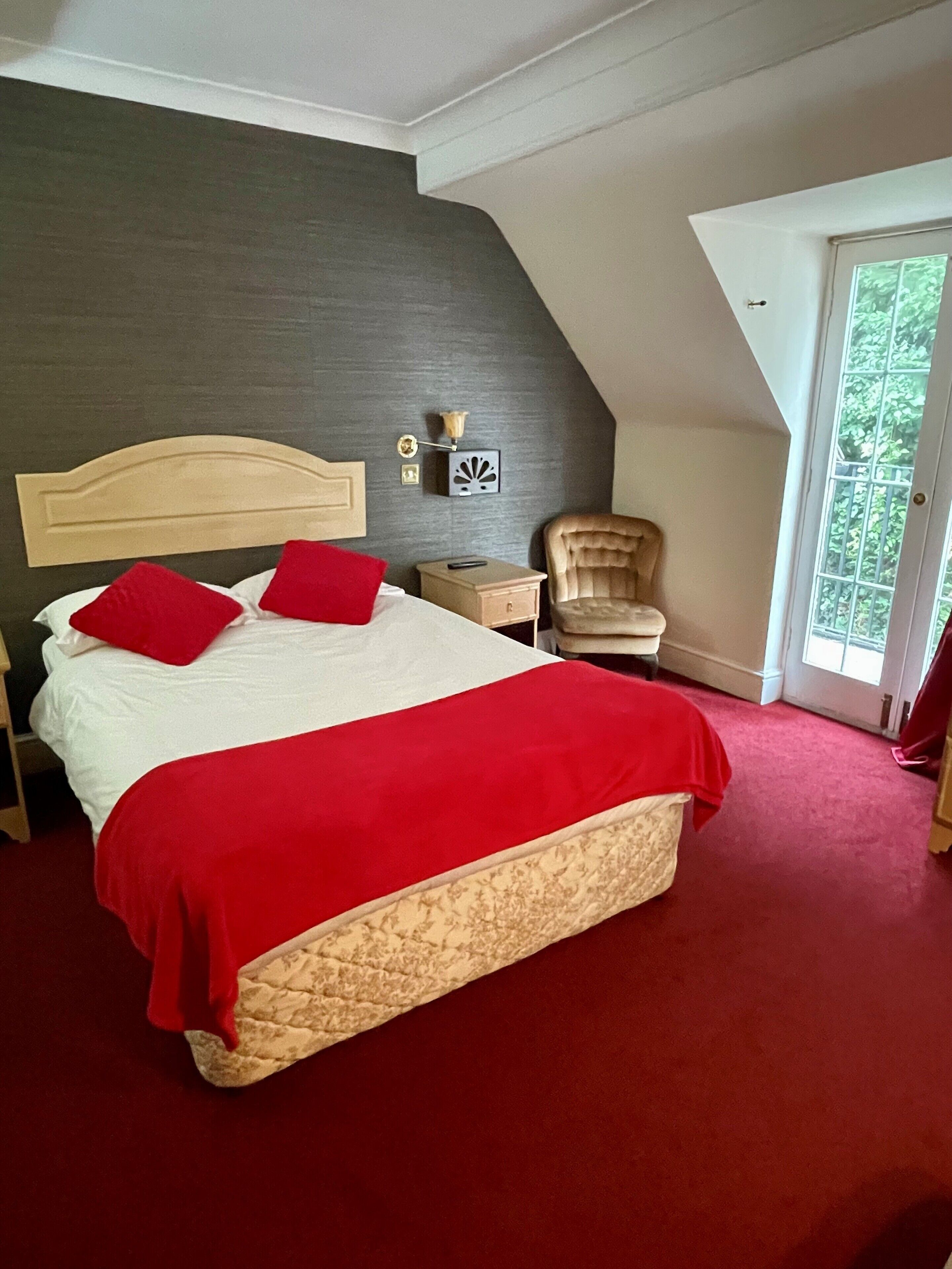 A bedroom with a large bed, red pillows, a nightstand, a chair, and a view of greenery through a door.