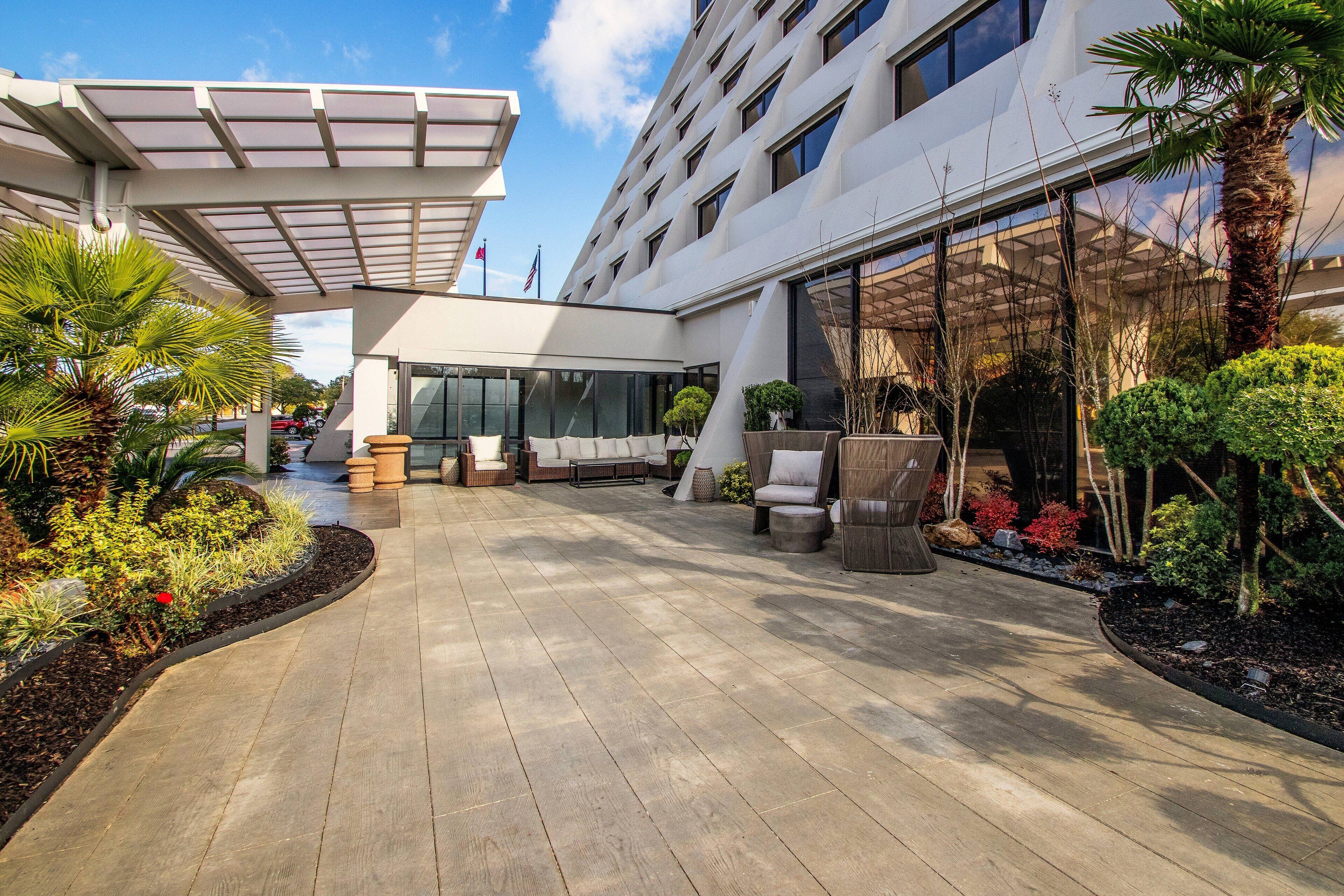 A modern hotel lobby with a covered seating area, outdoor plants, and a pathway leading to the entrance.