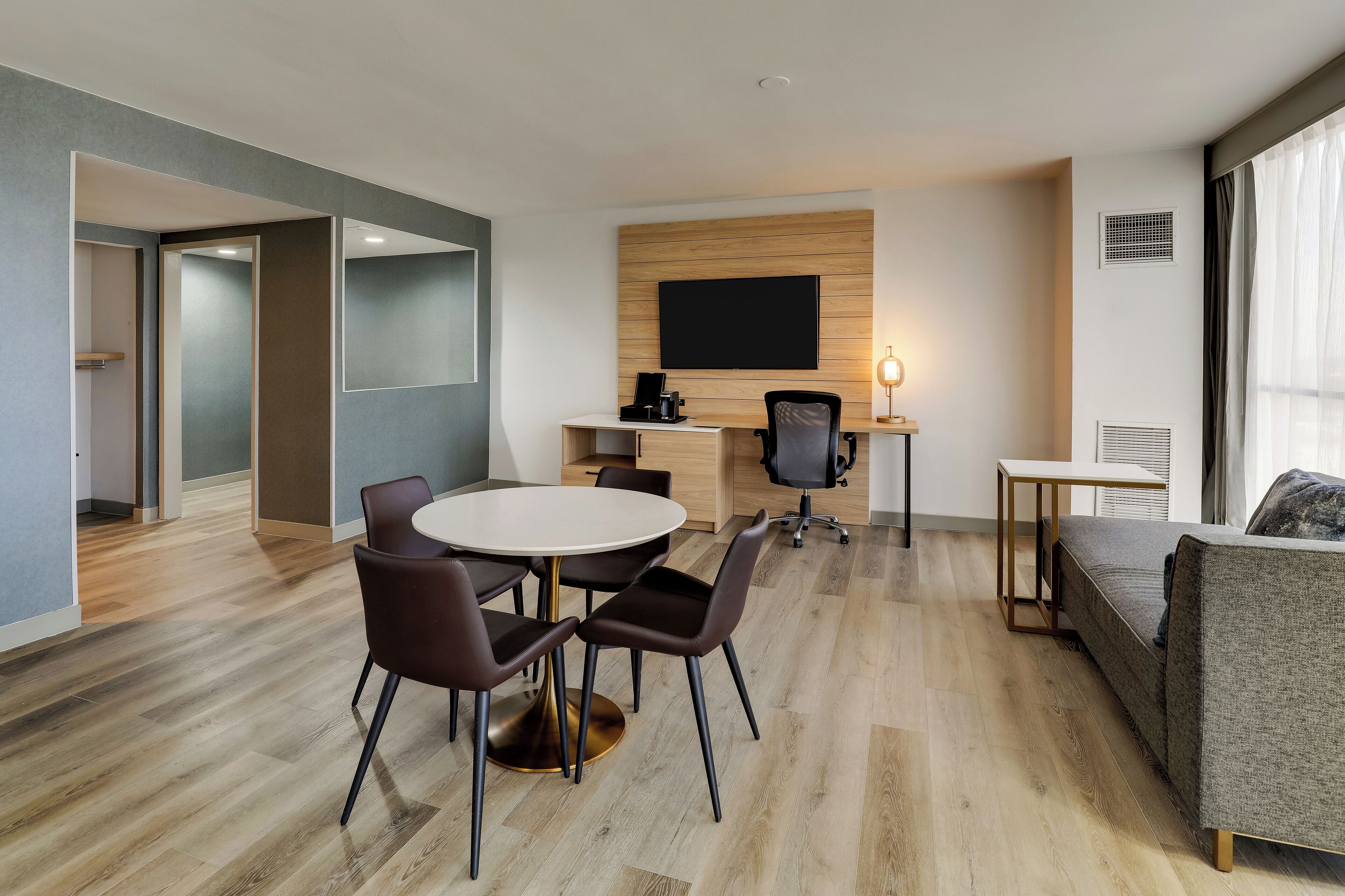 A modern hotel room with a dining area, a sofa, a TV mounted on the wall, and a desk with a chair.
