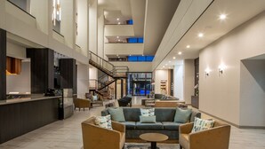 A spacious lobby with a high ceiling, a central seating area with sofas and armchairs, a reception desk, and a staircase leading to upper floors. - Mobile Marriott (Mobile)