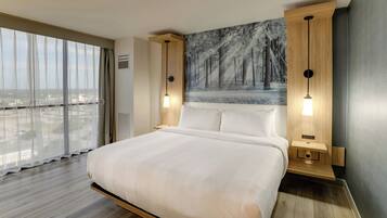 A modern hotel room with a large bed, wooden headboard, and a city view through floor-to-ceiling windows.