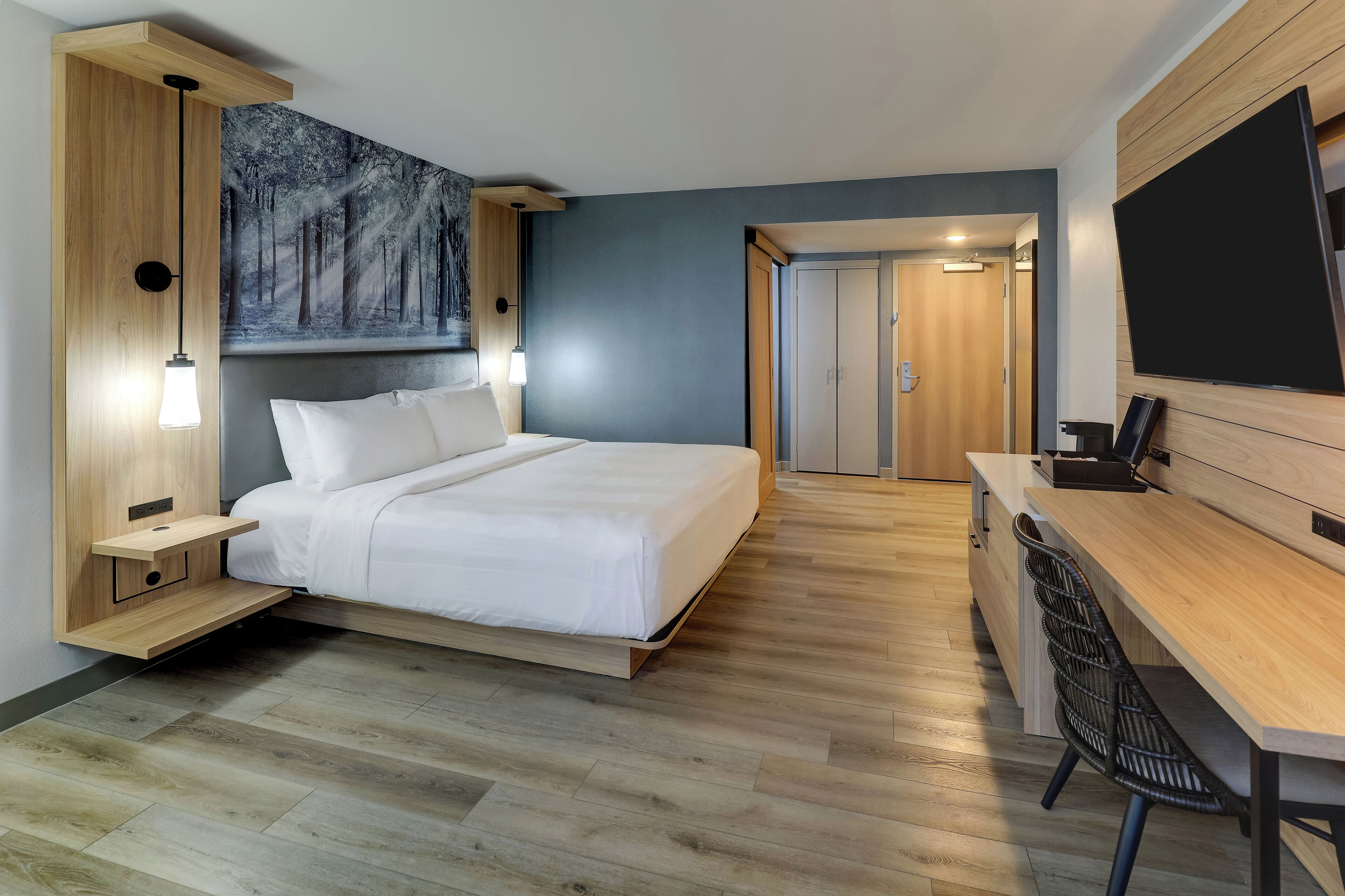 A modern hotel room with a bed, bedside tables, a desk, and a large TV.