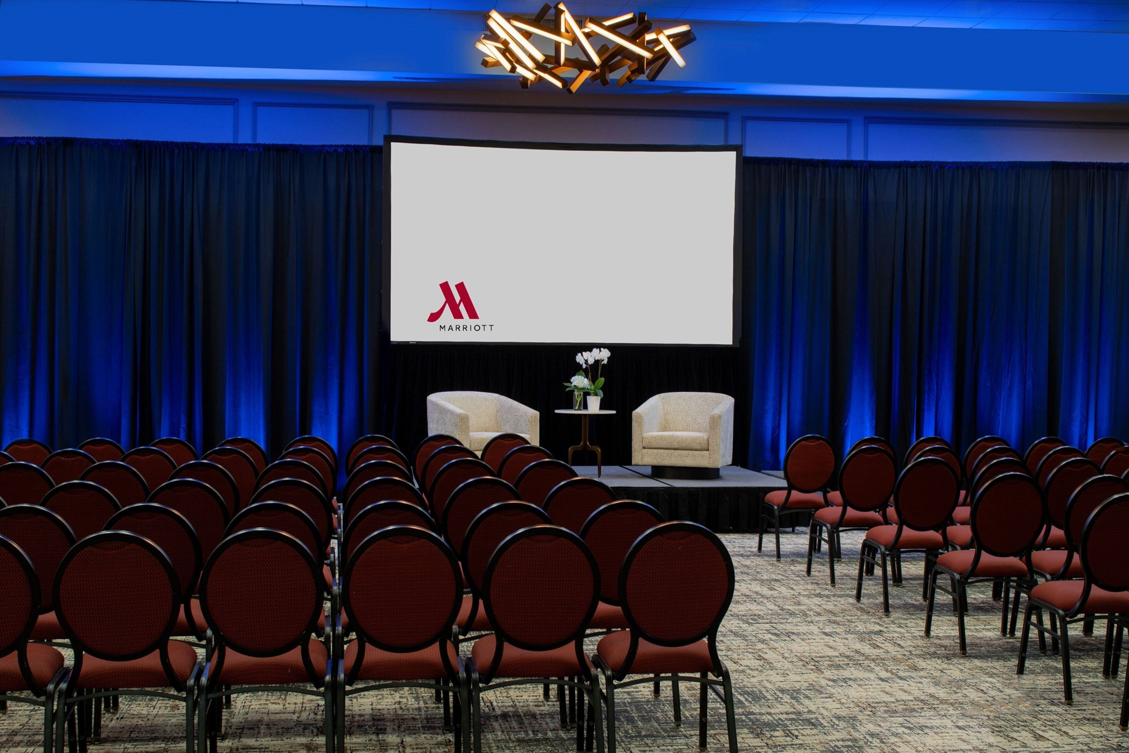 Un salón de conferencias con filas de sillas rojas, un escenario con una pantalla grande mostrando el logotipo de Marriott y dos sillones.