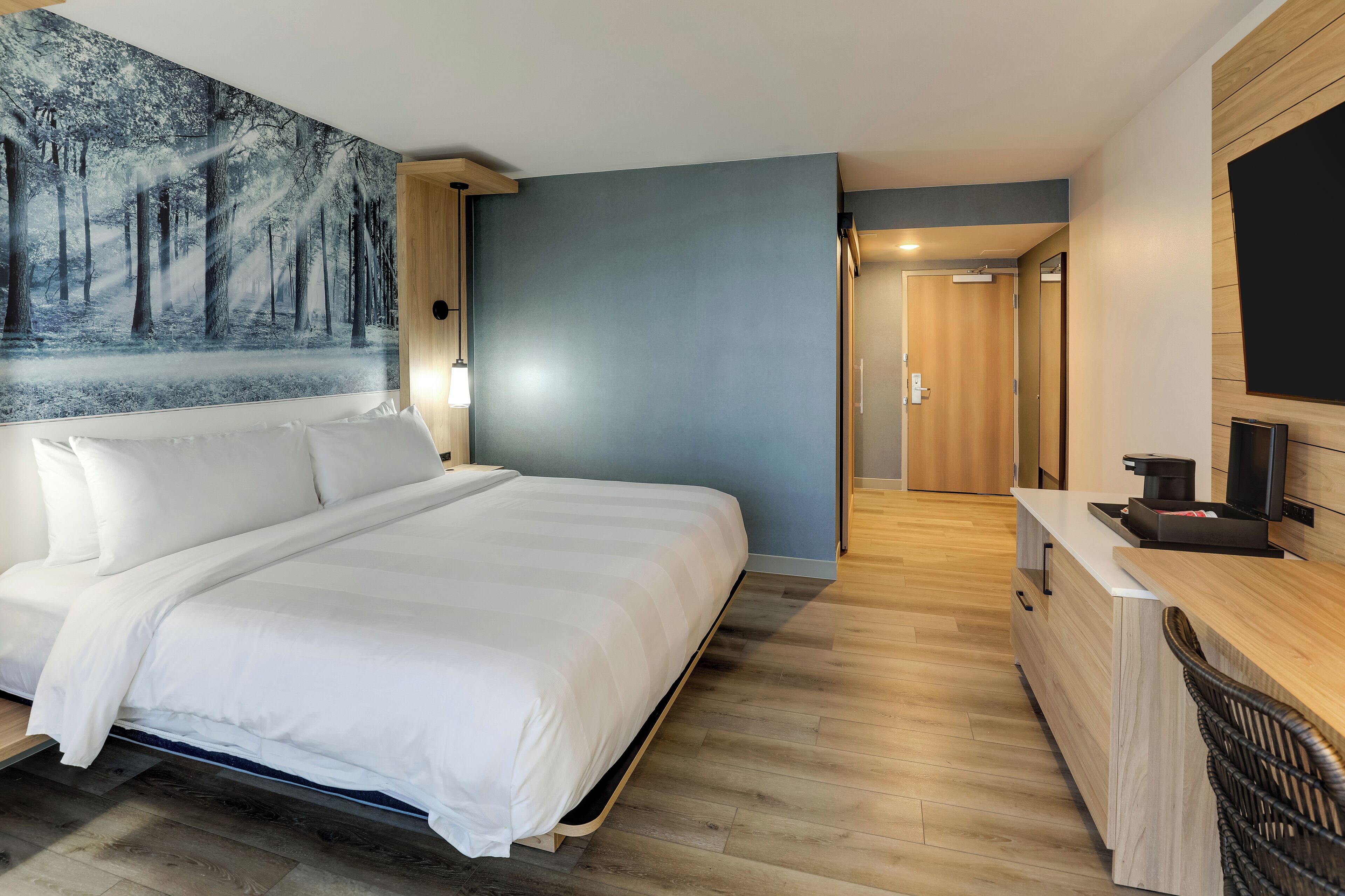 A modern hotel room with a large bed, a painting of a forest, a wooden desk, and a television.