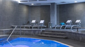 Un área de piscina interior moderna con una piscina cristalina, escalones que bajan al agua y varias sillas de descanso con cojines.