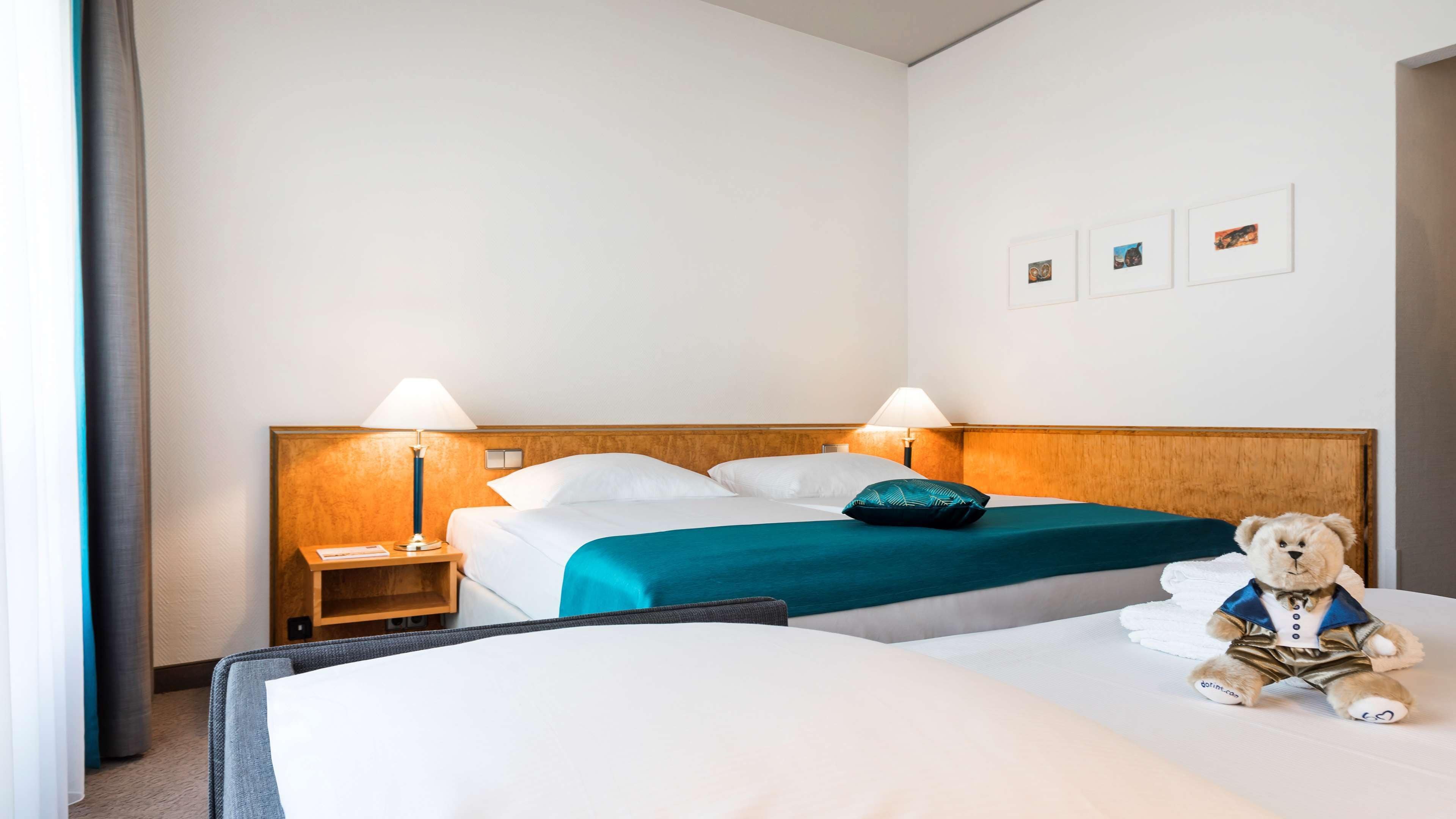 A hotel room with a double bed, two bedside lamps, a wooden headboard, a teal bedspread, and a teddy bear on the bed.