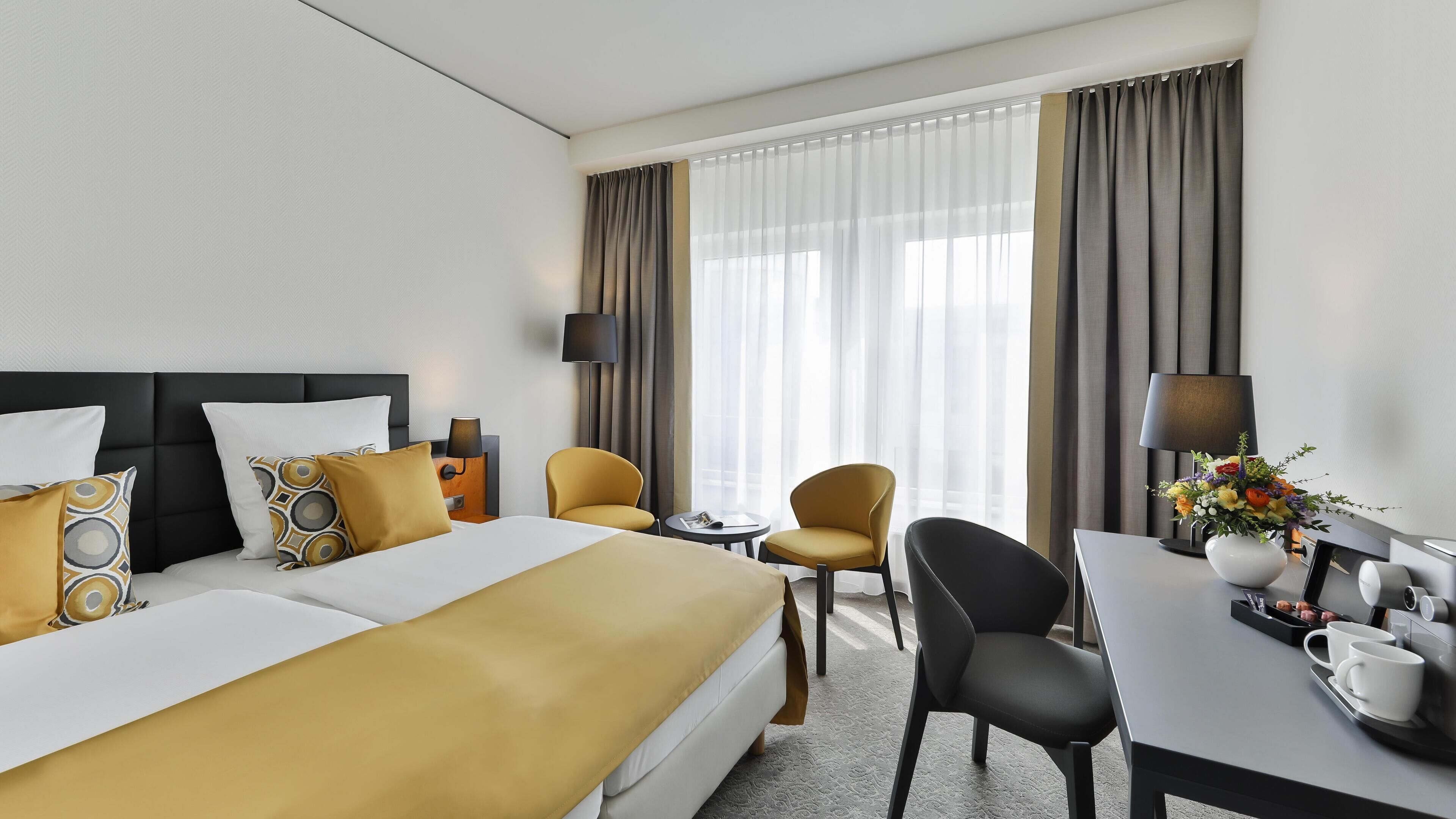 Ein Hotelzimmer mit Bett, zwei Stühlen, einem kleinen Tisch, einer Kaffeemaschine und einem Fenster mit Vorhängen.