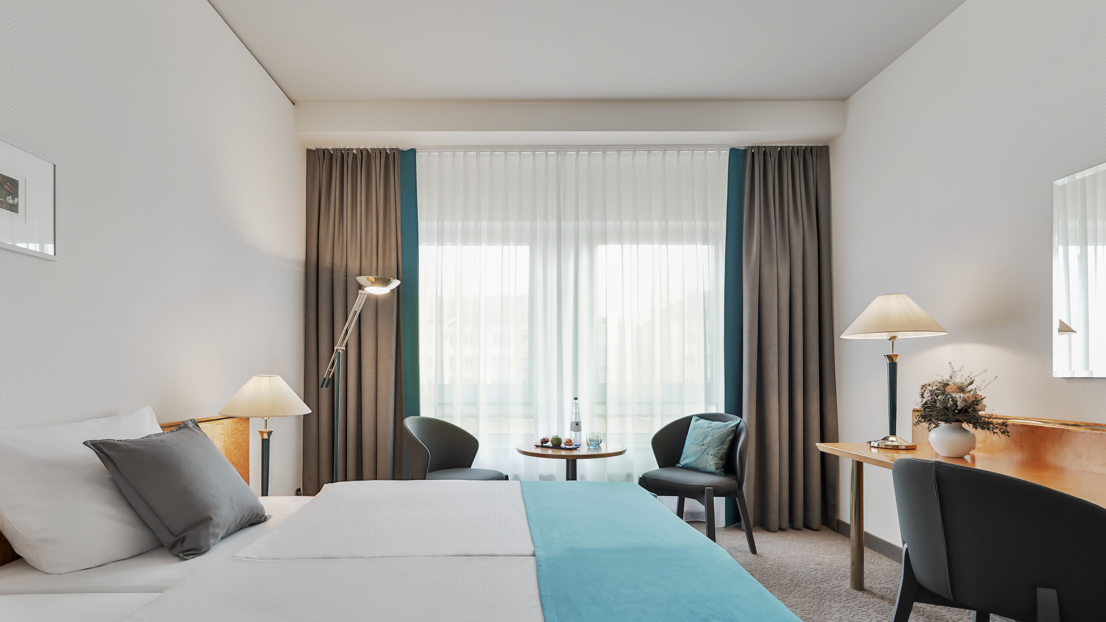 Una habitación de hotel con cama, dos sillas, un escritorio, una lámpara y una ventana con cortinas.
