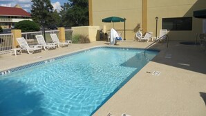 A swimming pool with lounge chairs and umbrellas, surrounded by a fence and a building with a red roof. - Fairview Inn and Suites (Mobile)