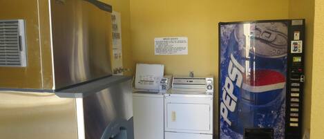 Une pièce équipée d’un réfrigérateur en acier inoxydable, d’un lave-linge, d’un sèche-linge et d’un distributeur automatique de boissons Pepsi.