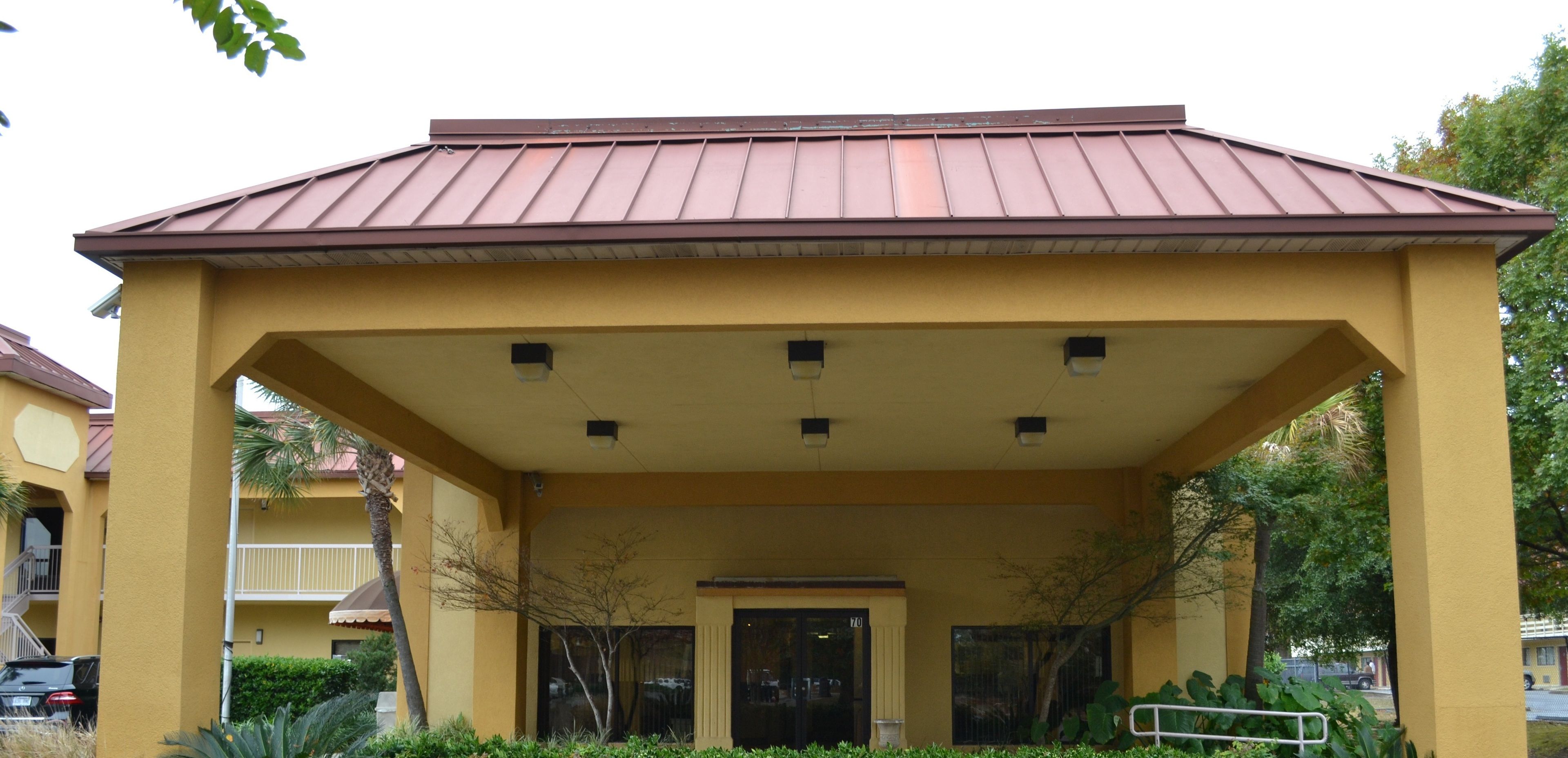 A yellow building with a brown roof, a covered entrance, and a small garden area with plants and a bench.