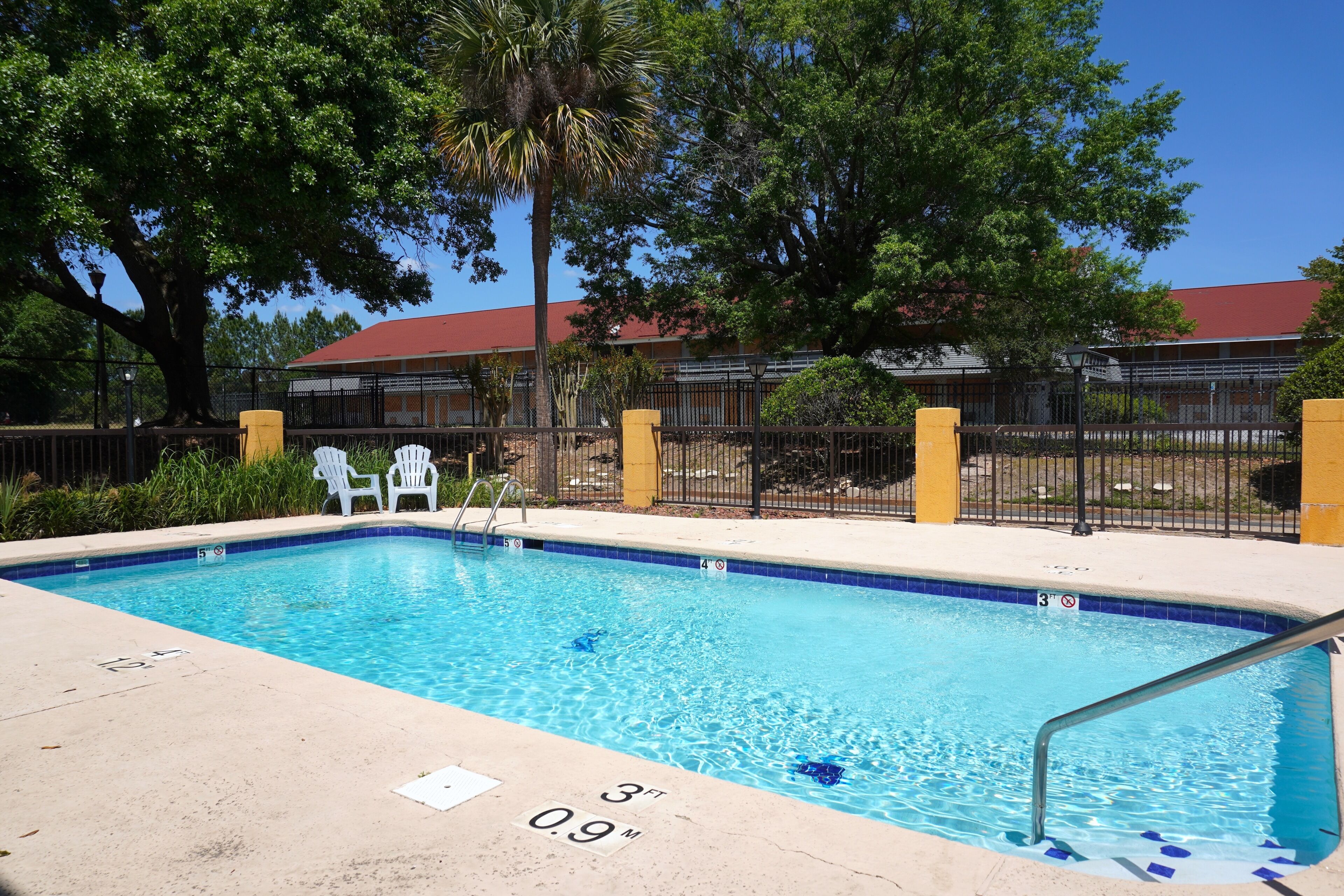 Una piscina con agua cristalina azul, rodeada por una valla y árboles, con un edificio al fondo.