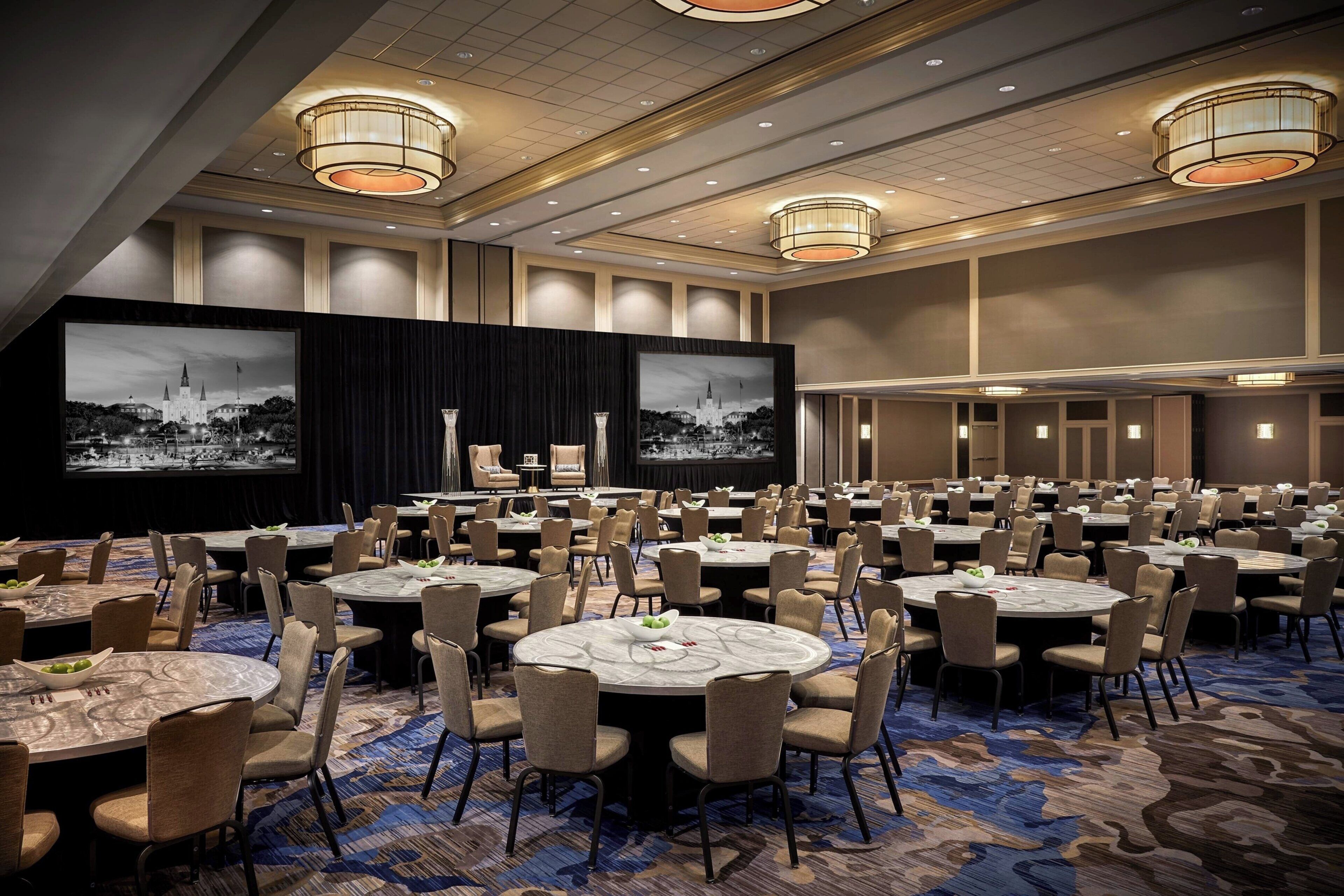 Une grande salle de conférence équipée de tables rondes et de chaises, d’une scène avec un décor en fond et de grands écrans diffusant des images.