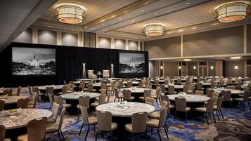 Une grande salle de conférence équipée de tables rondes et de chaises, d’une scène avec un décor en fond et de grands écrans diffusant des images.