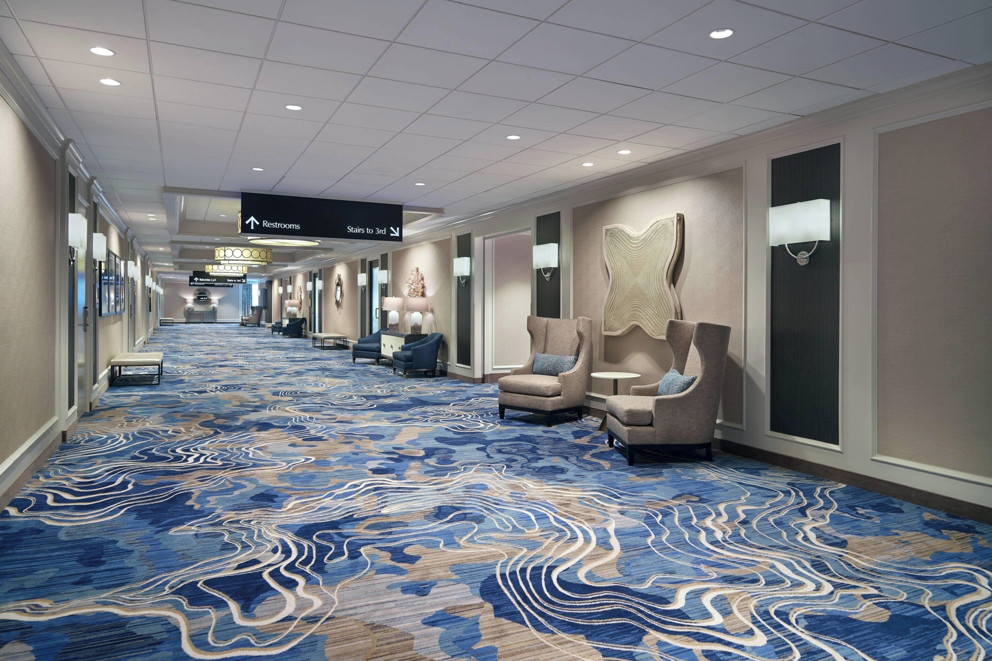 A hotel corridor with a blue patterned carpet, armchairs, and directional signs to restrooms and stairs.