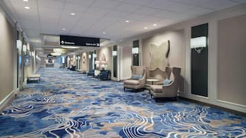 Corredor de hotel com carpete estampado em azul, poltronas e placas indicando banheiros e escadas.