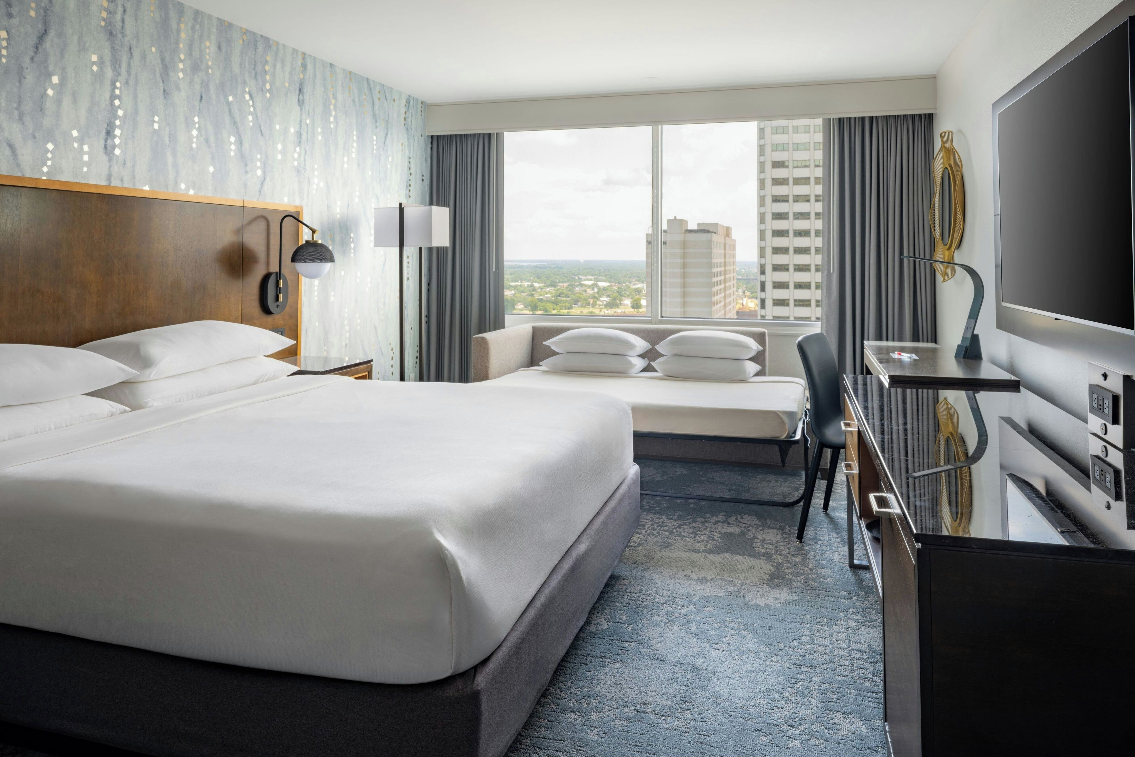 A hotel room with a large bed, a desk with a flat-screen TV, a chair, and a view of the cityscape.