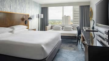 A hotel room with a large bed, a desk with a flat-screen TV, a chair, and a view of the cityscape.