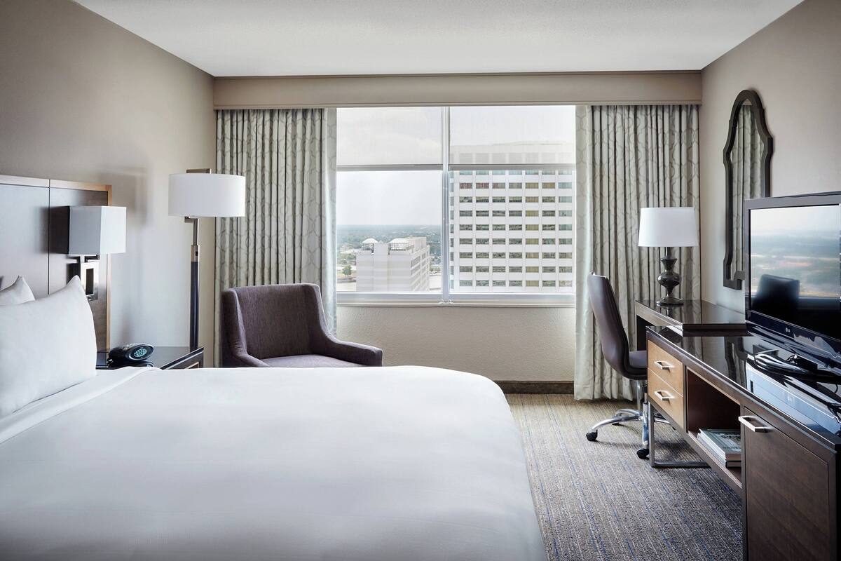 A hotel room with a large bed, a desk with a TV, a chair, a lamp, and a view of a cityscape through the window.