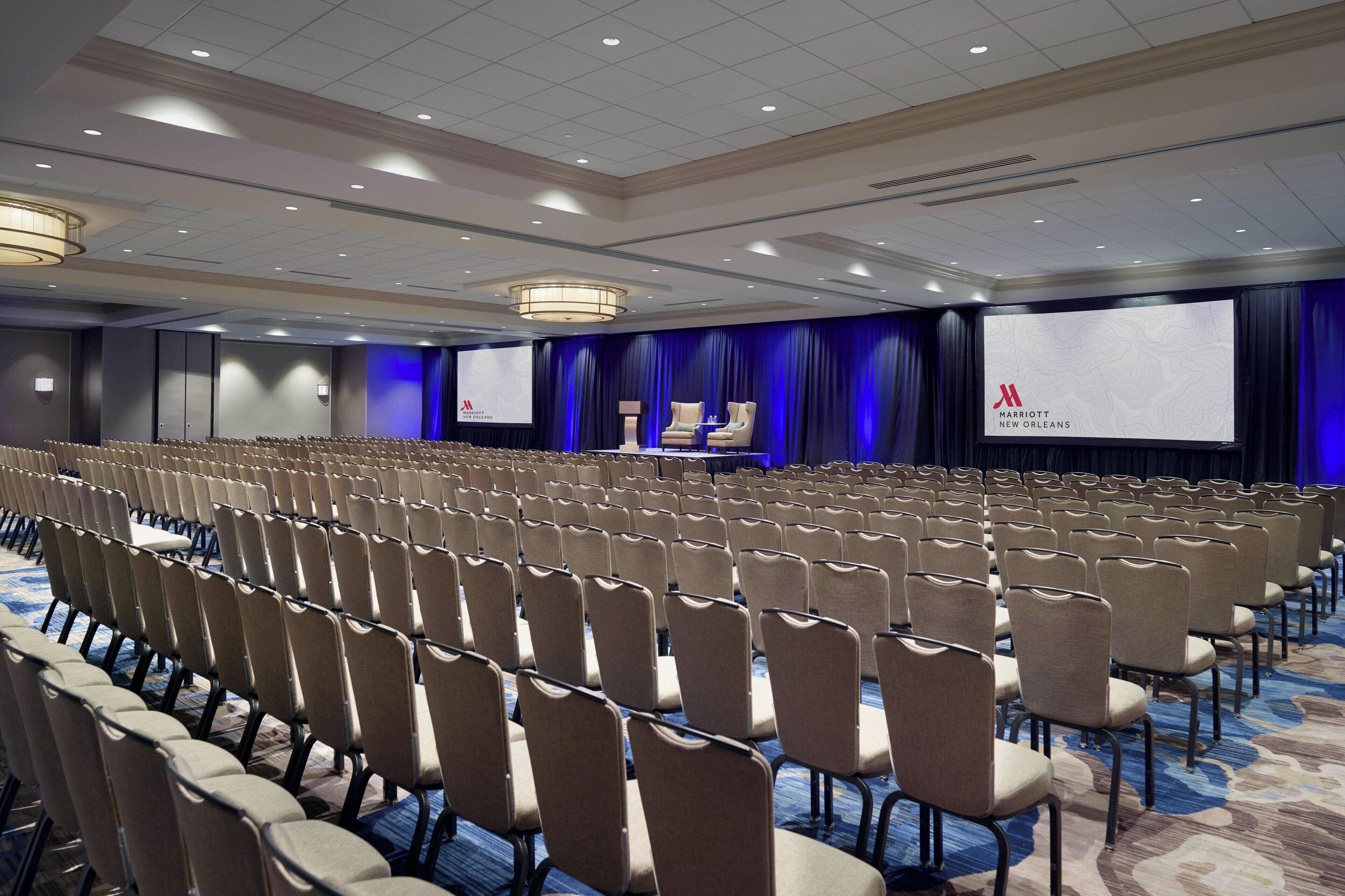 Un salón de conferencias con filas de sillas, un escenario con una pantalla y un telón de fondo azul.