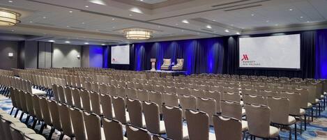 Un salón de conferencias con filas de sillas, un escenario con una pantalla y un telón de fondo azul.
