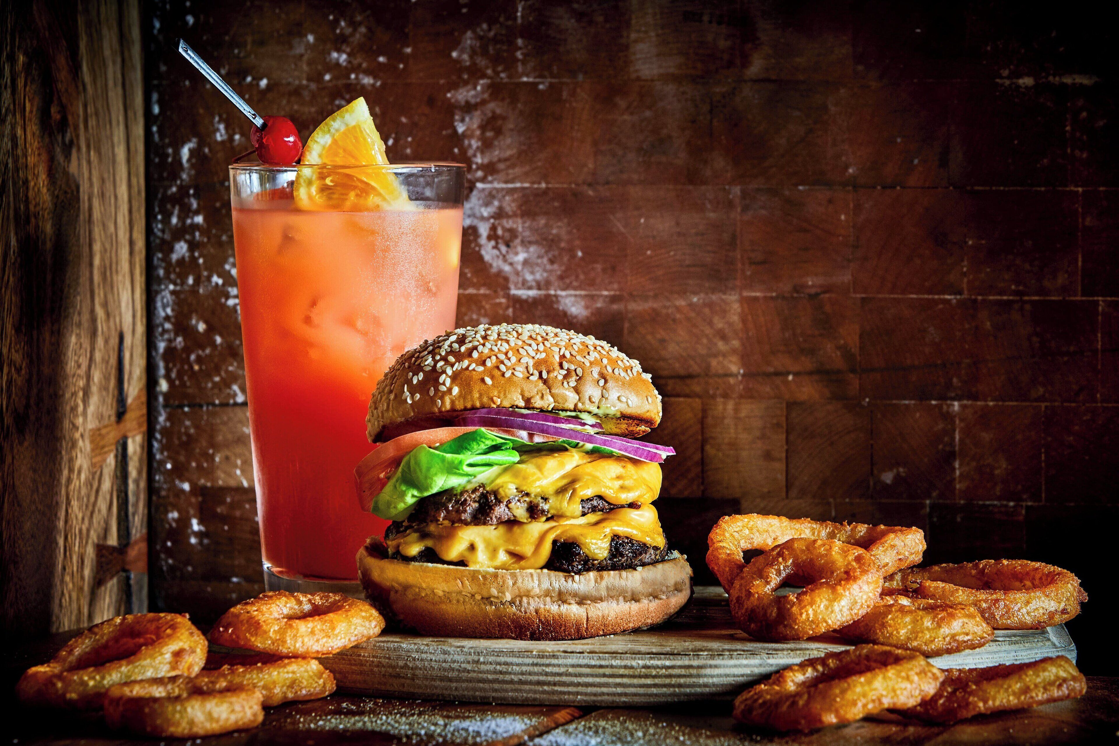 A burger with lettuce, cheese, and a sesame seed bun, accompanied by onion rings and a glass of a red beverage with a cherry and lemon slice.