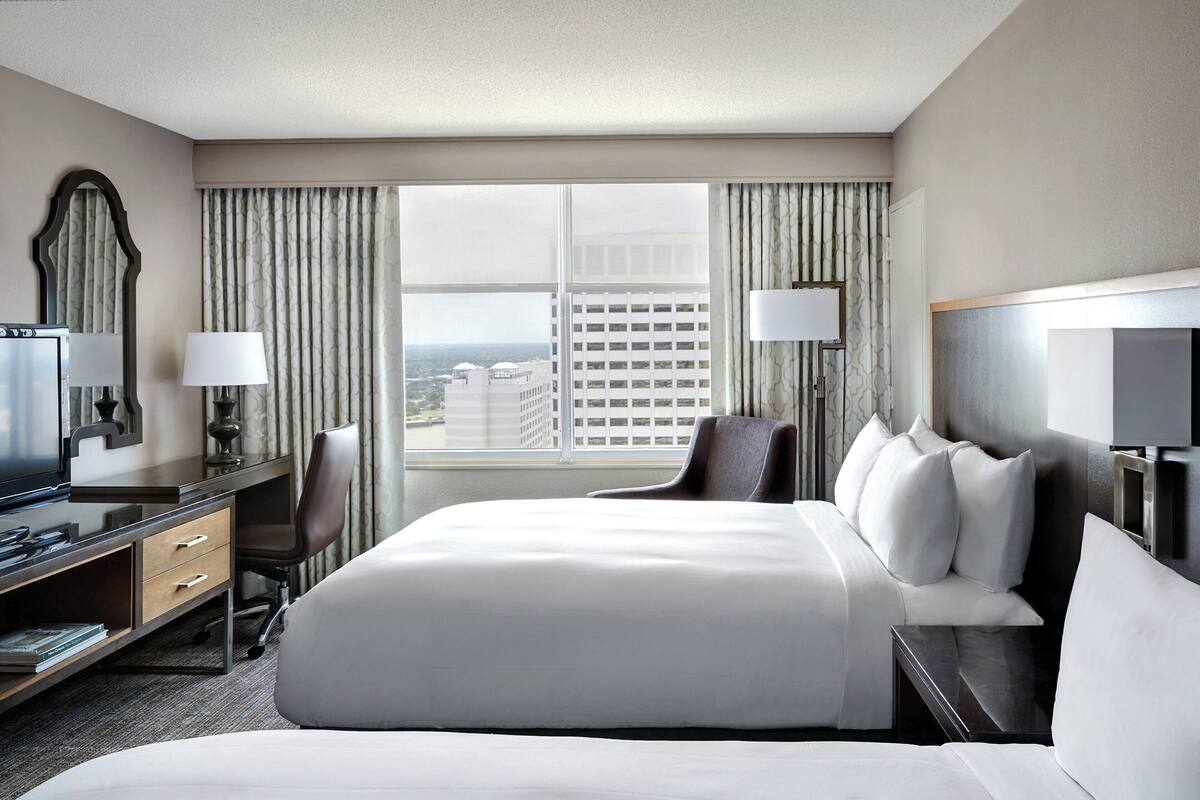 A hotel room with a large bed, a desk with a computer, a chair, and a view of the city through the window.