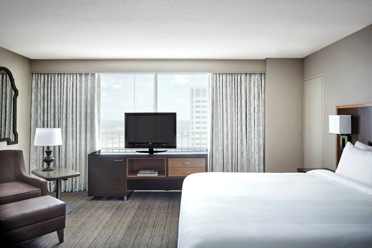 A hotel room with a bed, a TV on a stand, a desk with a lamp, a chair, and a view of the city through large windows.
