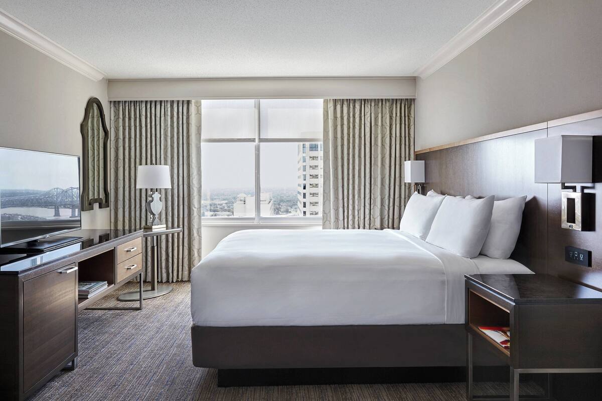 A hotel room with a large bed, a desk with a TV, a dresser, and a window with curtains.